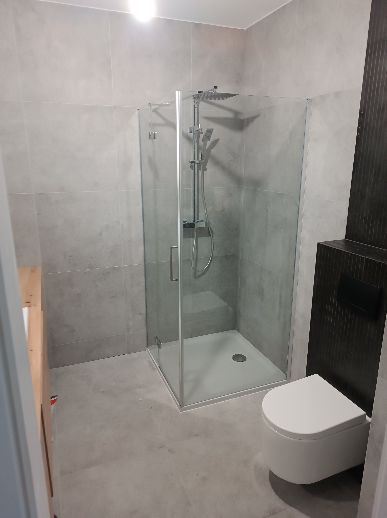 Nowoczesna łazienka z kabiną prysznicową typu walk-in, szare płytki na ścianach i podłodze, wisząca toaleta i ciemny panel dekoracyjny z ukrytym systemem spłukiwania.