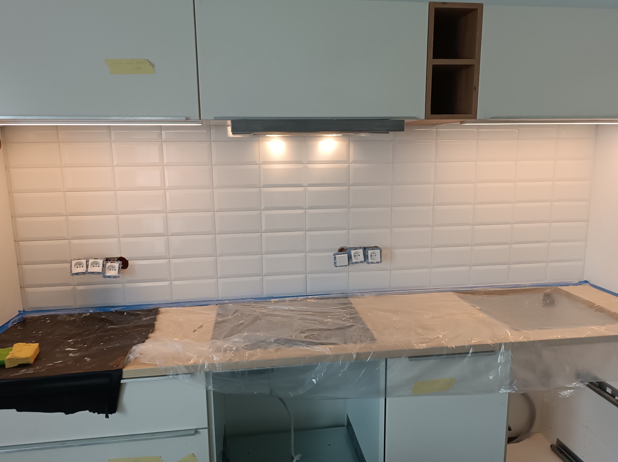 Białe płytki metra na ścianie kuchni, z zainstalowanymi gniazdkami elektrycznymi i okapem kuchennym, blat zabezpieczony folią ochronną, widoczne szafki kuchenne.