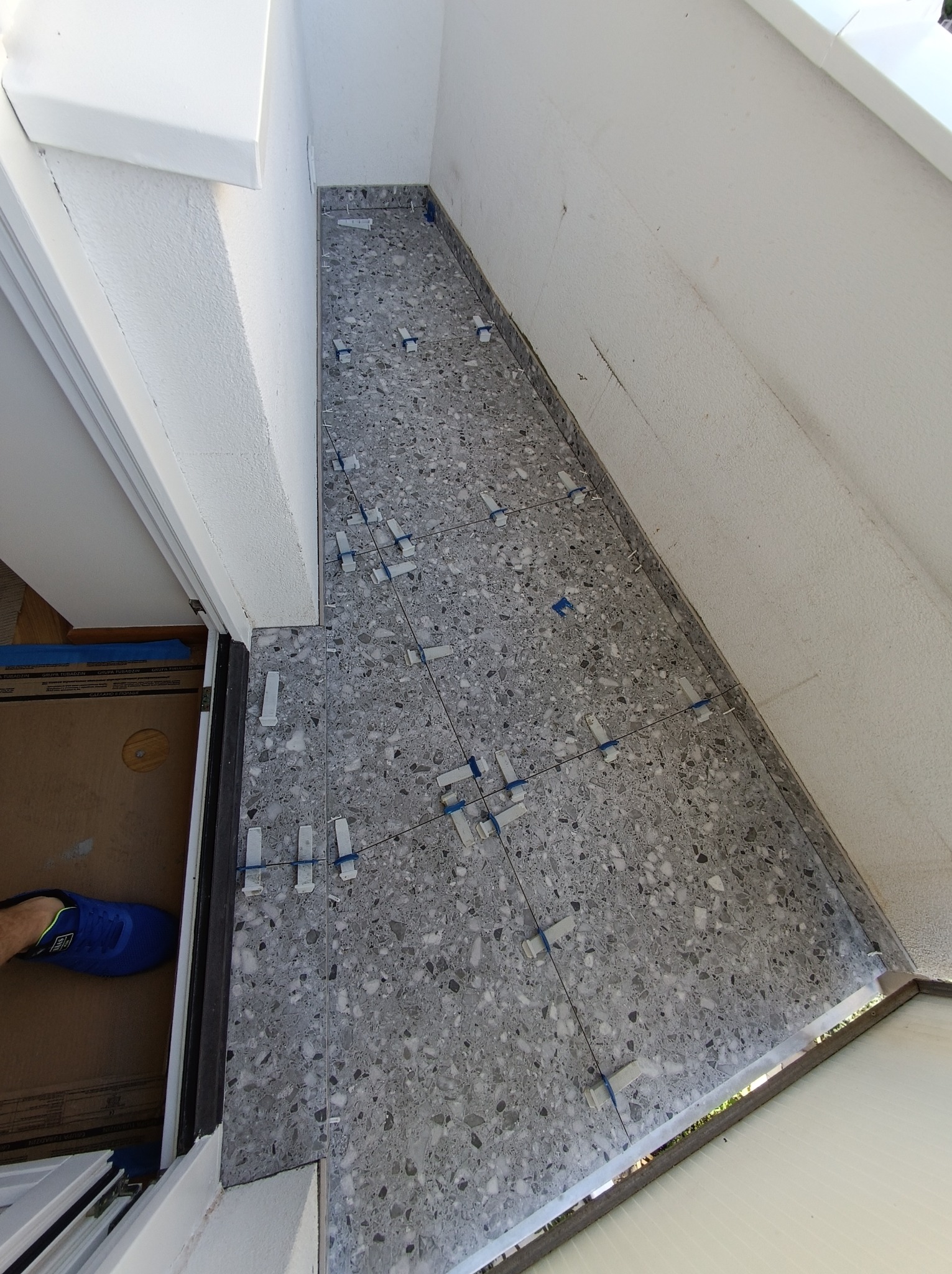 Szara, nakrapiana terakota ułożona na podłodze, z użyciem poziomujących klipsów i sznurka, widoczny fragment buta pracownika i fragment drzwi.