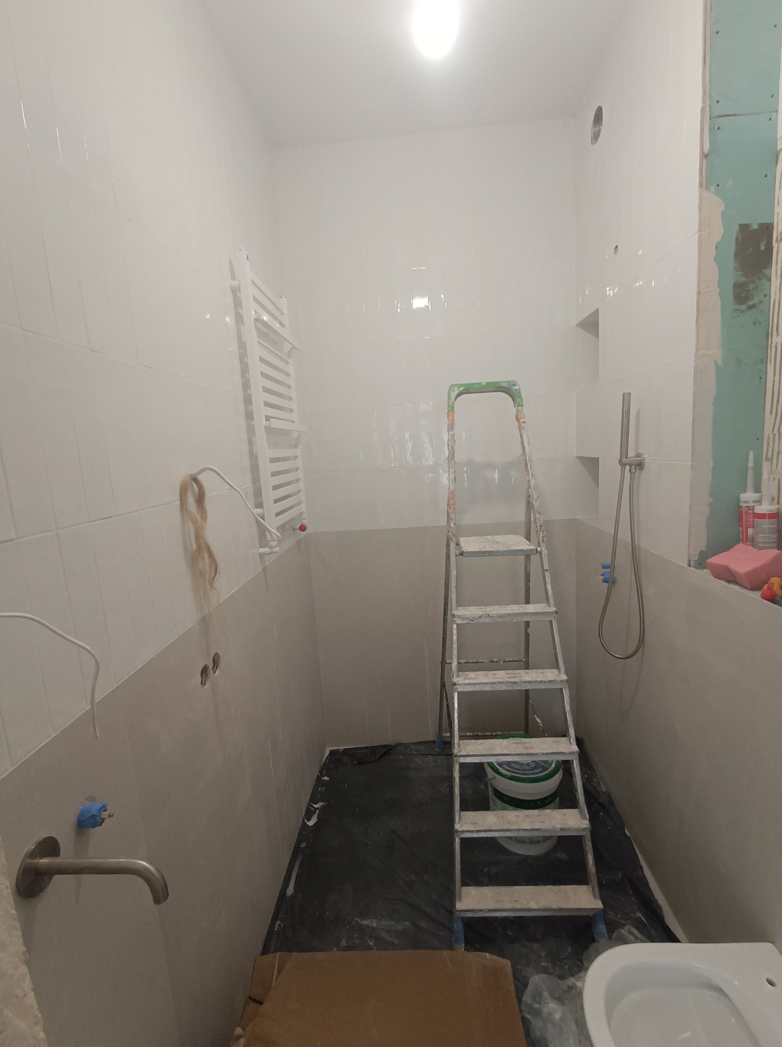Wnętrze łazienki w trakcie prac wykończeniowych: białe płytki na ścianach, drabina malarska, częściowo zamontowana toaleta, widoczne instalacje hydrauliczne i fragment ściany z płyt...