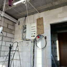 Patryk Falkowski - Wnętrze w trakcie prac elektrycznych: rozdzielnia elektryczna, kable prowadzone wzdłuż ściany z betonu komórkowego, drabina i rury instalacyjne na podłodze.