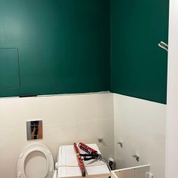 Patryk Falkowski - Remont łazienki: ściana pomalowana na ciemnozielono nad białymi płytkami, widoczna toaleta, pralka z poziomicą i tubą silikonu, oraz szafka pod umywalkę w trakcie montażu.