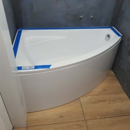 Patryk Falkowski - Nowa, narożna wanna akrylowa zabezpieczona niebieską taśmą malarską, zamontowana w rogu łazienki z wyłożonymi płytkami imitującymi drewno i szarą hydroizolacją na ścianach.