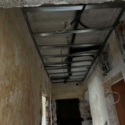 Patryk Falkowski - Metalowa konstrukcja sufitu podwieszanego w trakcie montażu, widoczne przewody elektryczne, surowe ściany z cegły i resztki starego tynku.