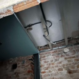 Patryk Falkowski - Sufit podwieszany w trakcie montażu, widoczne profile metalowe, instalacja elektryczna i częściowo zamontowana płyta gipsowo-kartonowa na tle surowej, ceglanej ściany z widocznymi ubytkami tynku.