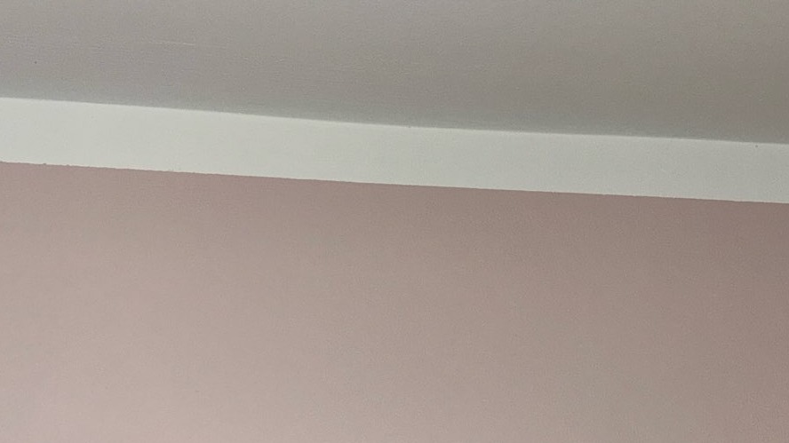 Fragment ściany pomalowanej na delikatny róż z białą listwą przysufitową i jasnym sufitem. Widoczny delikatny odcień światła na suficie.