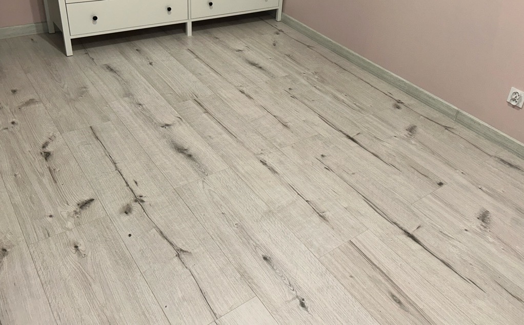 Jasne panele podłogowe z wyraźnym wzorem drewna ułożone w nowym pokoju z różową ścianą i białą listwą przypodłogową, widoczny fragment białej komody.