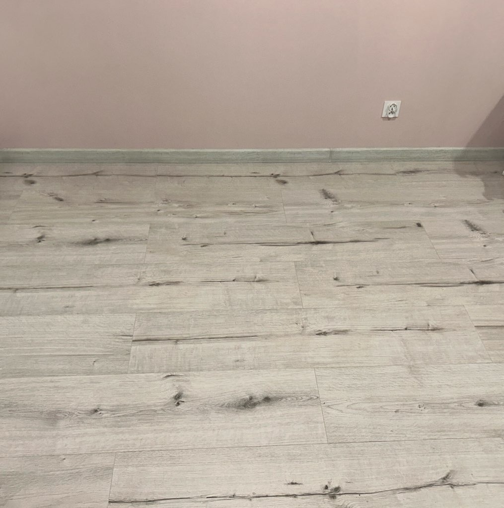 Jasne panele podłogowe z widoczną strukturą drewna, ułożone na podłodze przy różowej ścianie z listwą przypodłogową i gniazdkiem elektrycznym.