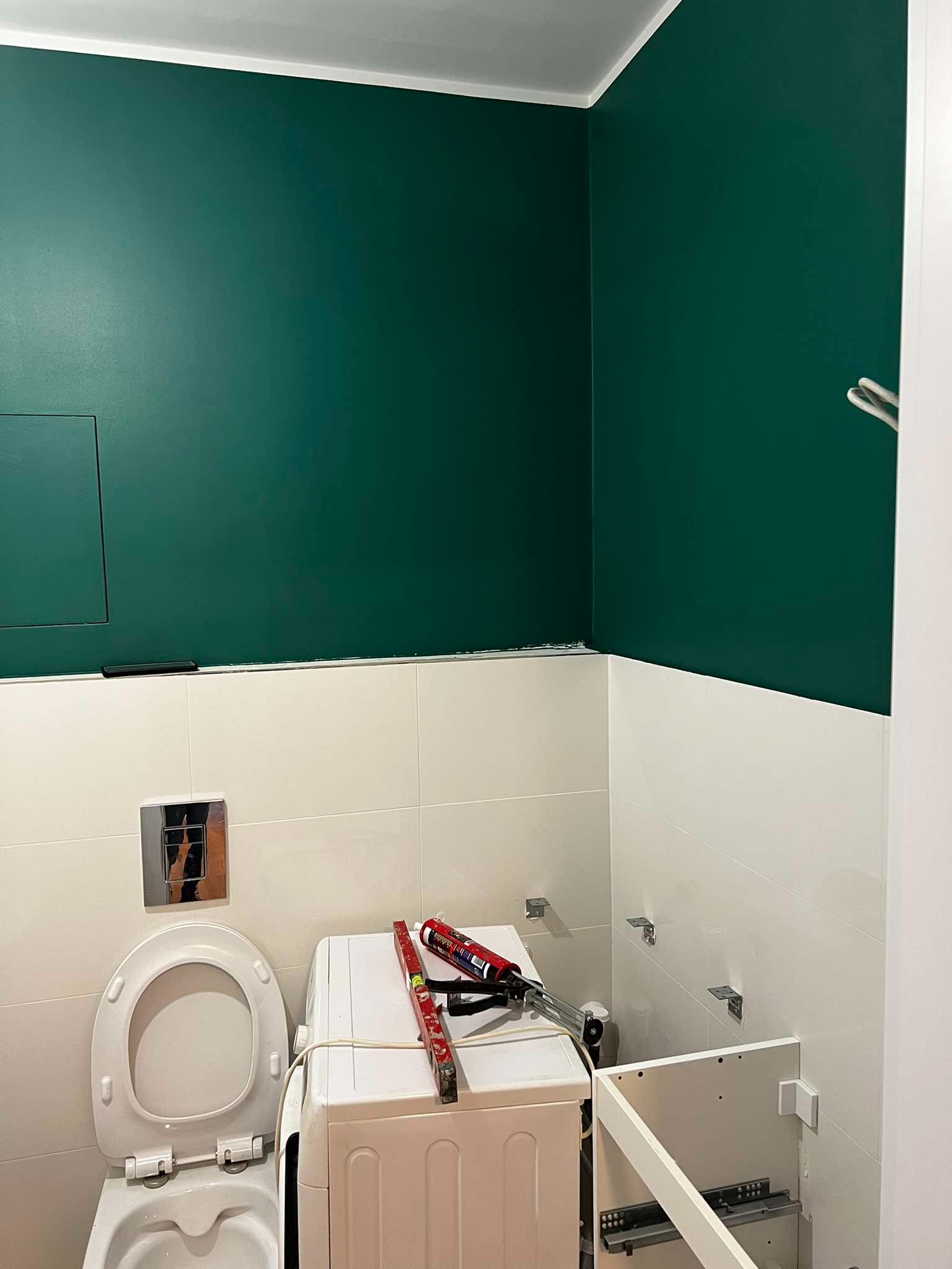 Remont łazienki: ściana pomalowana na ciemnozielono nad białymi płytkami, widoczna toaleta, pralka z poziomicą i tubą silikonu, oraz szafka pod umywalkę w trakcie montażu.