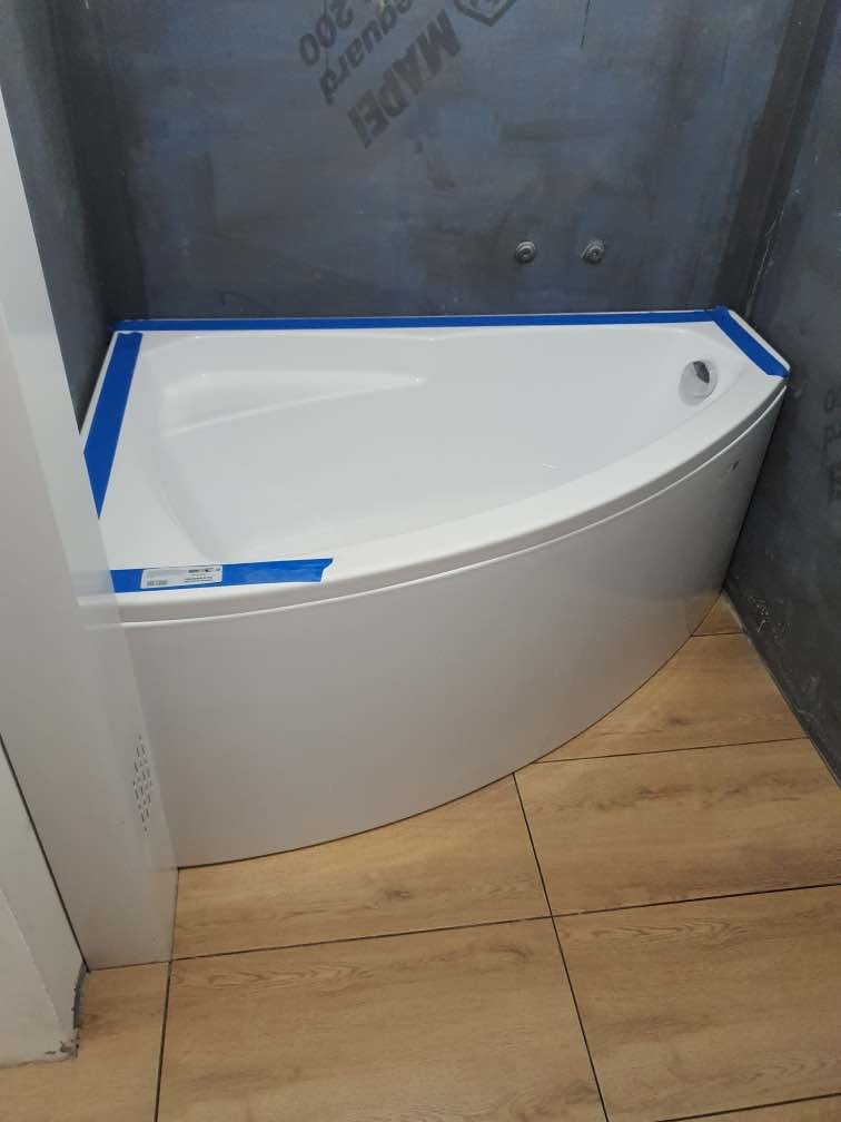 Nowa, narożna wanna akrylowa zabezpieczona niebieską taśmą malarską, zamontowana w rogu łazienki z wyłożonymi płytkami imitującymi drewno i szarą hydroizolacją na ścianach.