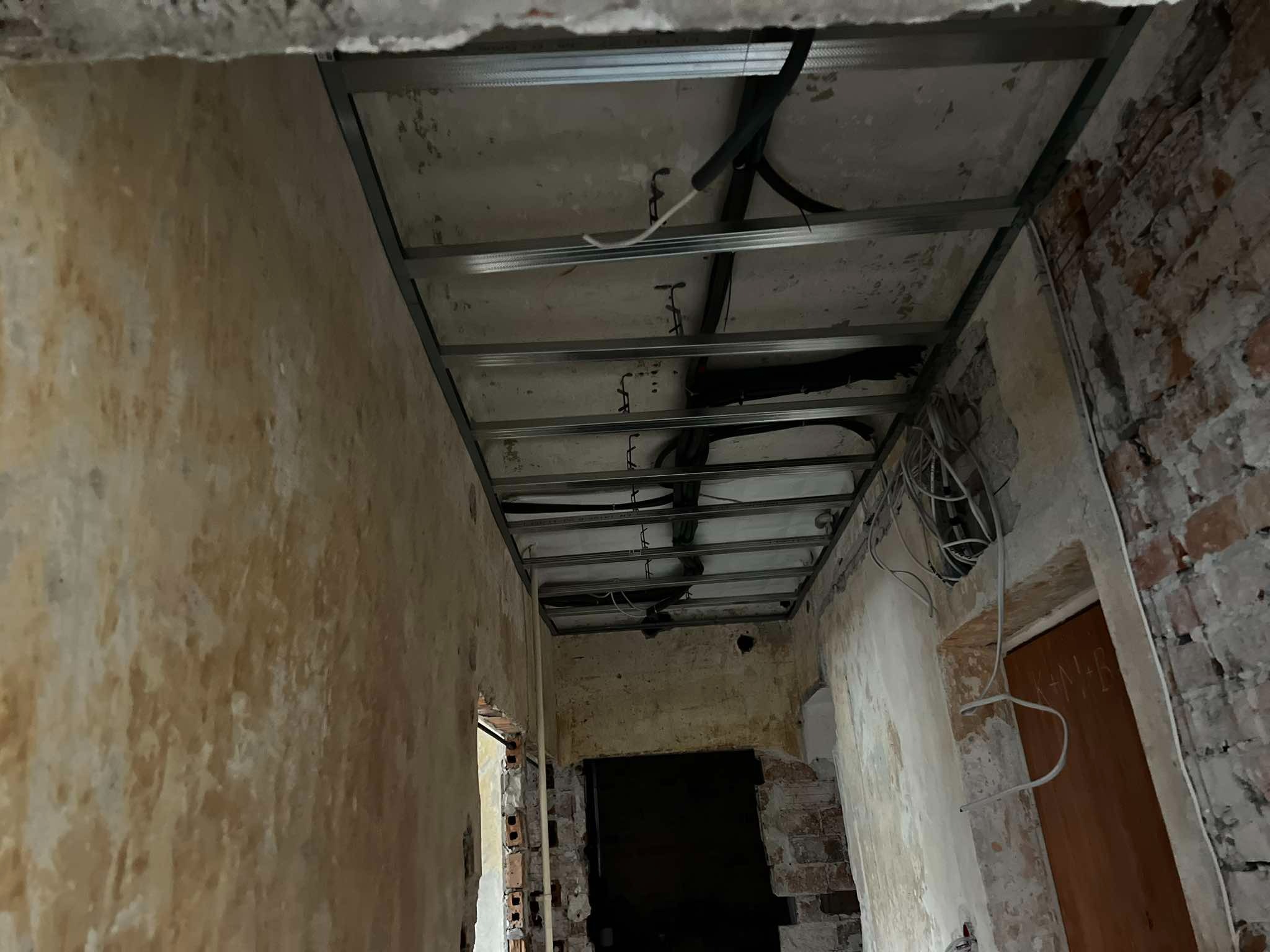 Metalowa konstrukcja sufitu podwieszanego w trakcie montażu, widoczne przewody elektryczne, surowe ściany z cegły i resztki starego tynku.