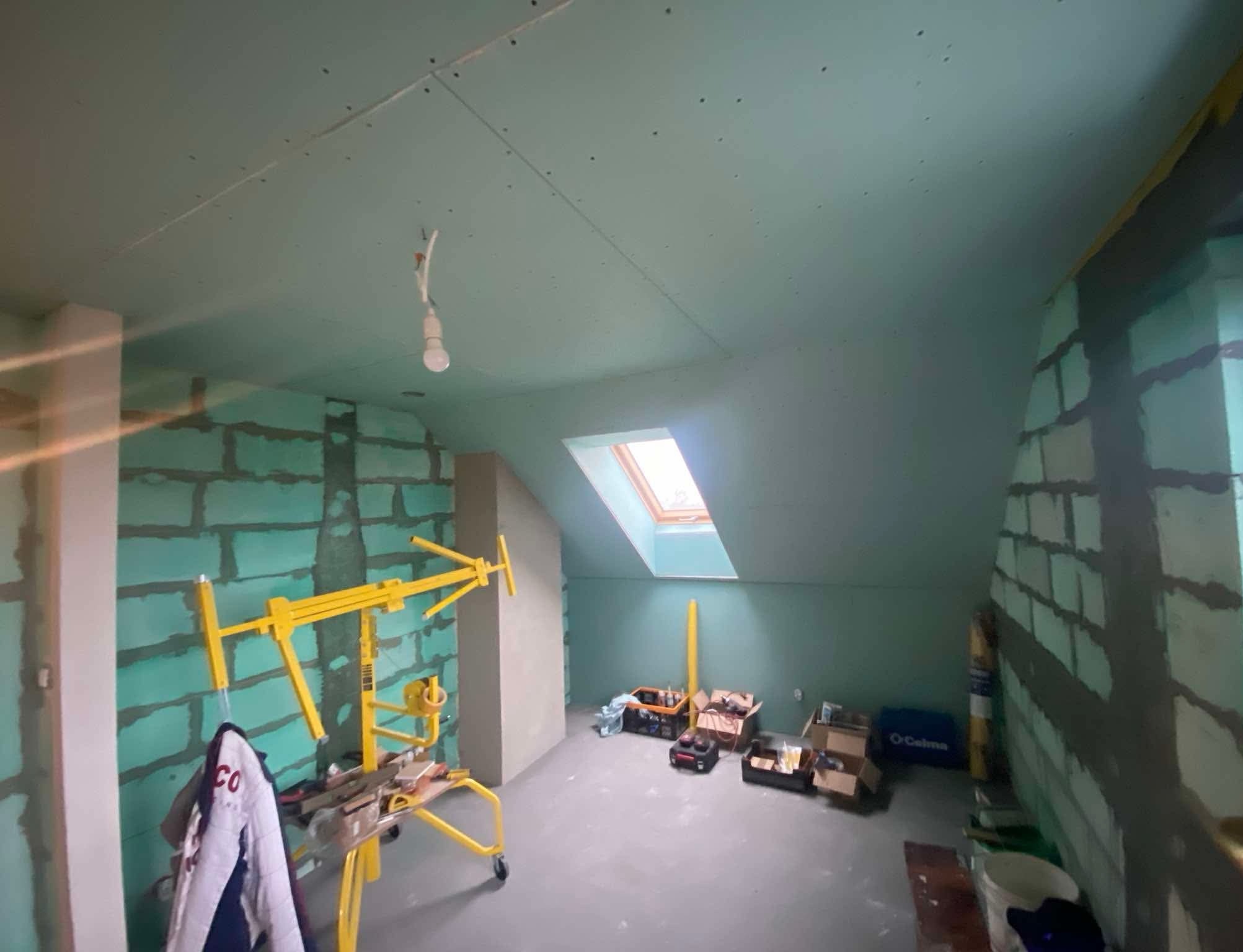 Poddasze w trakcie prac wykończeniowych: ściany częściowo pokryte płytami gipsowo-kartonowymi, widoczny podnośnik do płyt, narzędzia i materiały budowlane, okno dachowe wpadające do pomieszczenia...