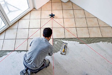 Mężczyzna w trakcie układania płytek podłogowych w pomieszczeniu ze skośnym sufitem, widoczne linie lasera wyznaczające kierunek, na podłodze rozprowadzony klej, obok kielnia.