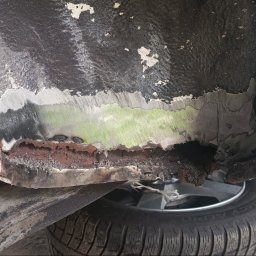 Bilard Garage - Fragment nadkola auta z widoczną korozją i śladami obróbki blacharskiej. Oczyszczony metal sąsiaduje z rdzawym nalotem. Widoczne opony samochodu.