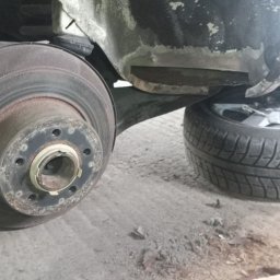 Bilard Garage - Błotnik samochodowy po naprawie blacharskiej, widoczna tarcza hamulcowa i opona. Ślady szpachli i spawania. Warsztatowy detal.