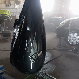 Bilard Garage - Błyszczący, czarny bak motocyklowy z logo, świeżo po lakierowaniu, zawieszony w warsztacie. W tle auto na podnośniku i narzędzia.