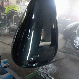 Bilard Garage - Błyszczący, ciemnozielony bak motocyklowy po lakierowaniu, zawieszony w warsztacie. Widoczne odbicia światła, fragment samochodu i narzędzia w tle.
