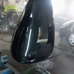 Błyszczący, ciemnozielony bak motocyklowy po lakierowaniu, zawieszony w warsztacie. Widoczne odbicia światła, fragment samochodu i narzędzia w tle.