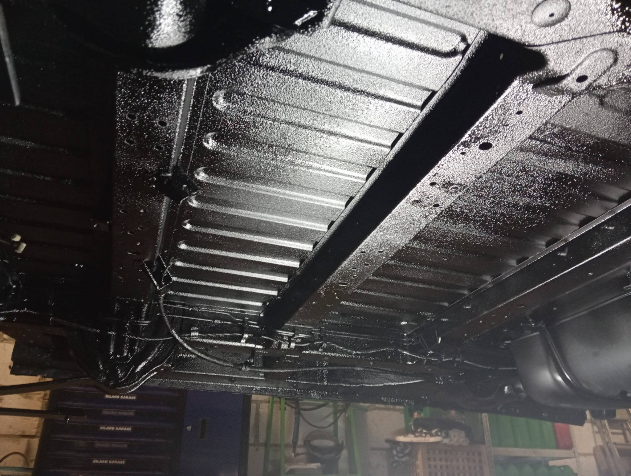 Podwozie samochodu po konserwacji, pokryte grubą warstwą czarnej, błyszczącej powłoki antykorozyjnej, widoczne elementy konstrukcyjne i przewody.