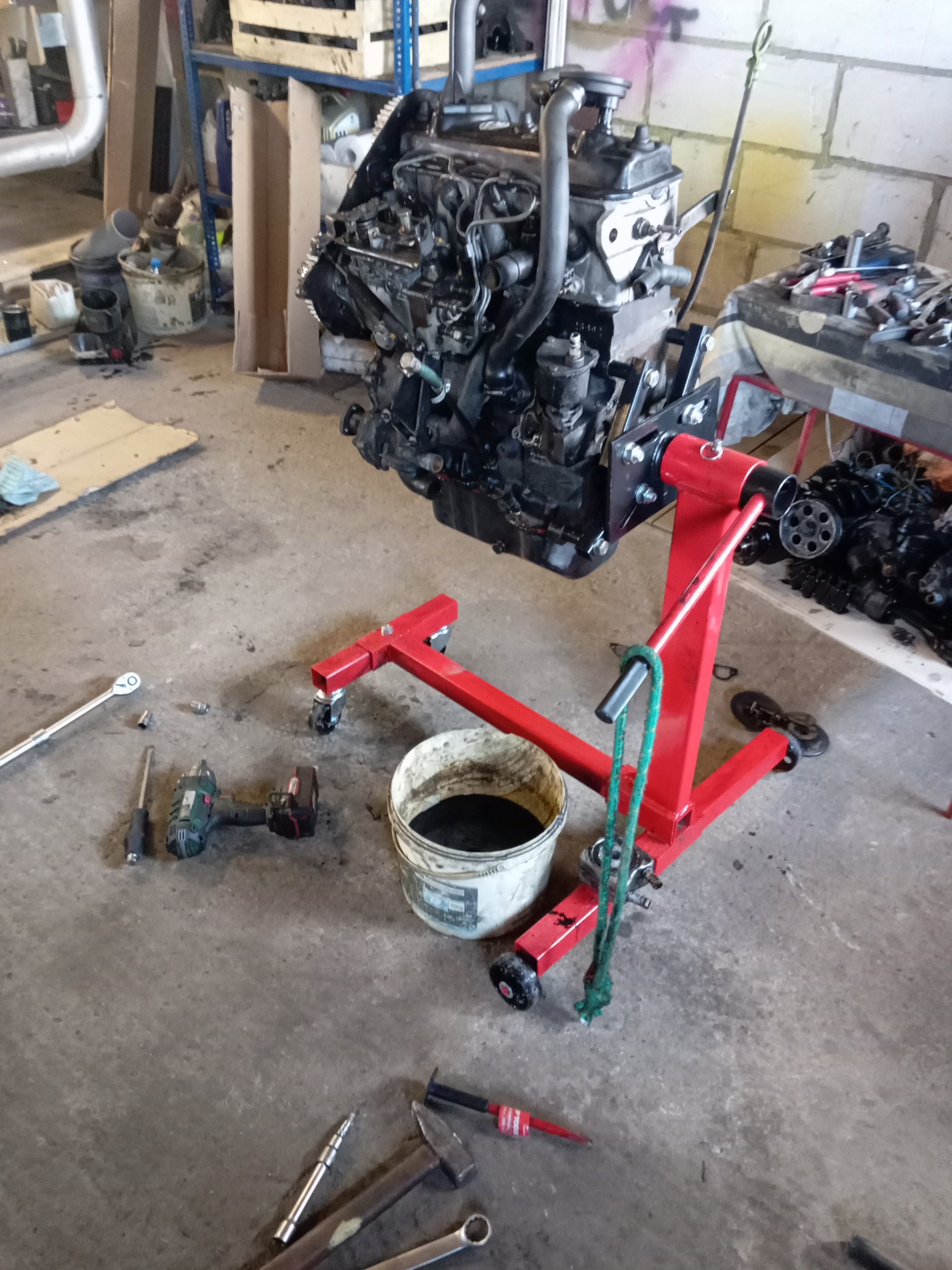Silnik samochodowy zawieszony na czerwonym stojaku warsztatowym, z widocznymi narzędziami i pojemnikiem na olej w tle warsztatu.