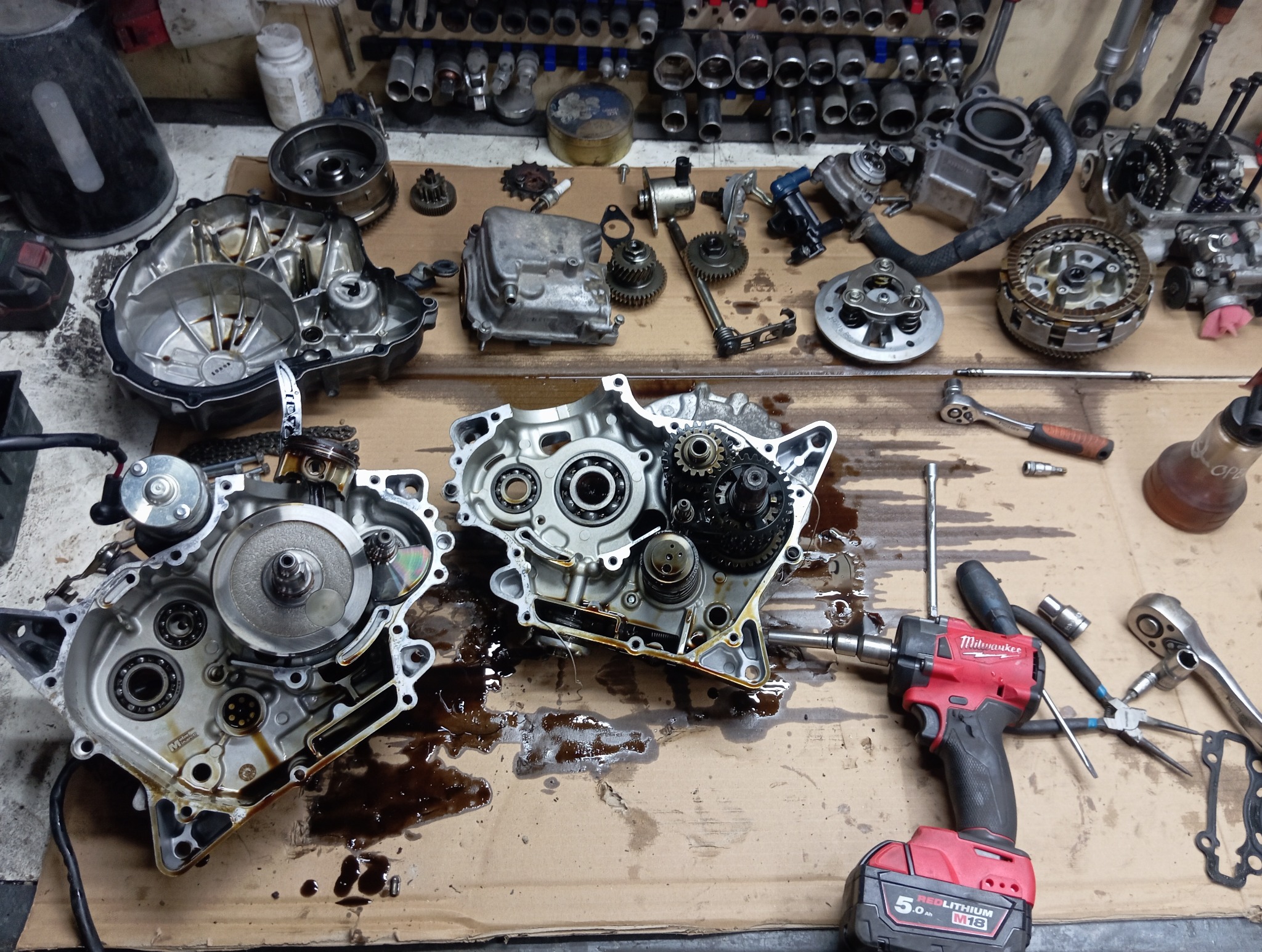 Rozłożony silnik motocyklowy na warsztatowym stole, widoczne narzędzia i części, w tym klucz udarowy Milwaukee. Detaliczny widok na proces naprawy.