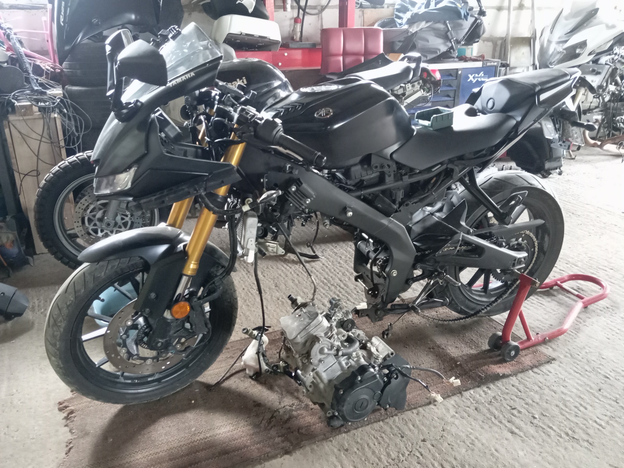 Czarny motocykl Yamaha bez owiewek, z widocznym silnikiem na podłodze warsztatu. Inne motocykle i opony w tle. Widoczne złote lagi zawieszenia.