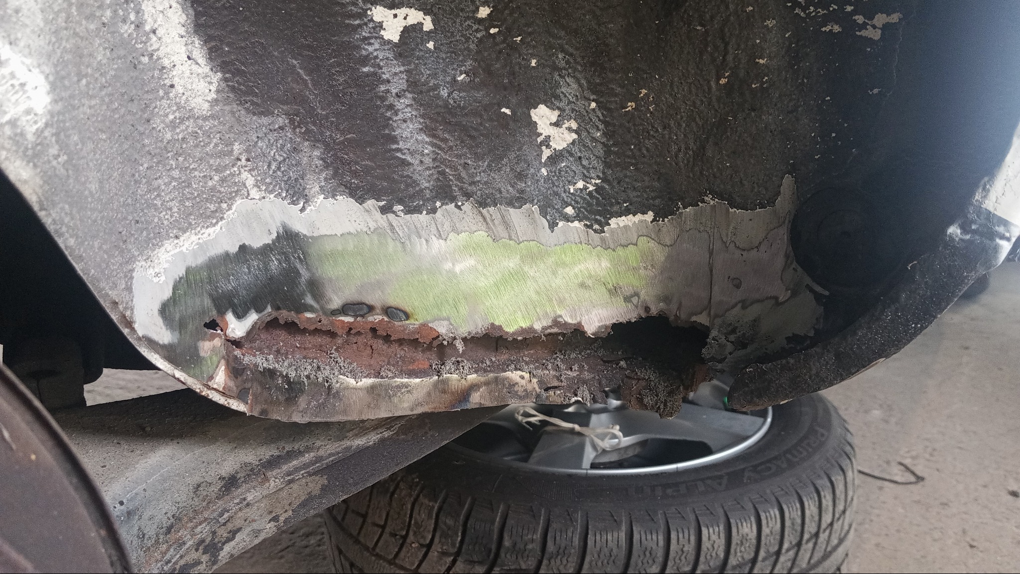 Fragment nadkola auta z widoczną korozją i śladami obróbki blacharskiej. Oczyszczony metal sąsiaduje z rdzawym nalotem. Widoczne opony samochodu.
