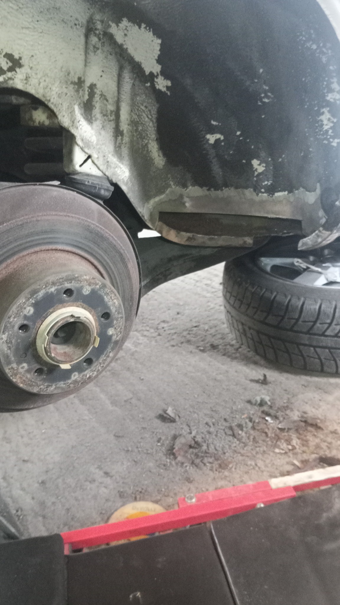 Błotnik samochodowy po naprawie blacharskiej, widoczna tarcza hamulcowa i opona. Ślady szpachli i spawania. Warsztatowy detal.