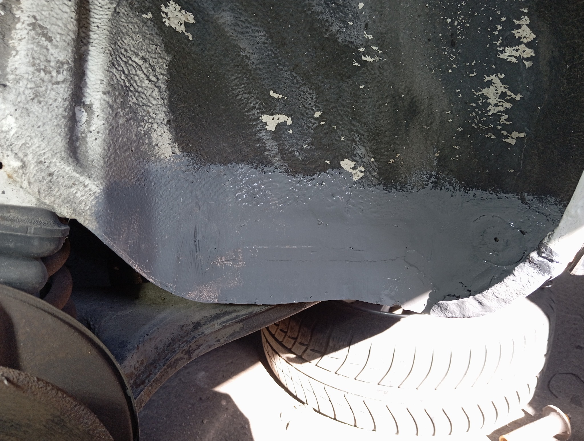 Wnętrze nadkola samochodu po zabezpieczeniu antykorozyjnym, z widocznymi śladami starej powłoki i nową, szarą warstwą ochronną. Widoczne opony i elementy zawieszenia.