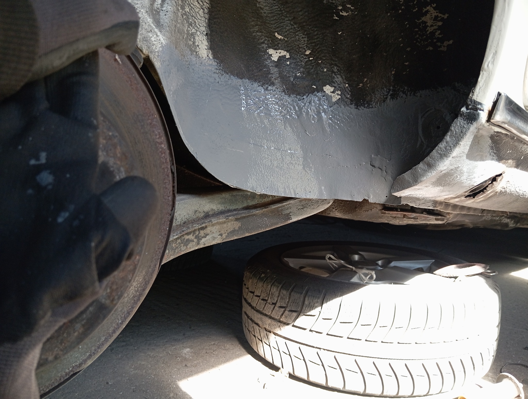 Wnętrze nadkola po zabezpieczeniu antykorozyjnym, widoczny element zawieszenia i opona. Szara, świeżo nałożona powłoka ochronna. Elementy metalowe z widoczną korozją.