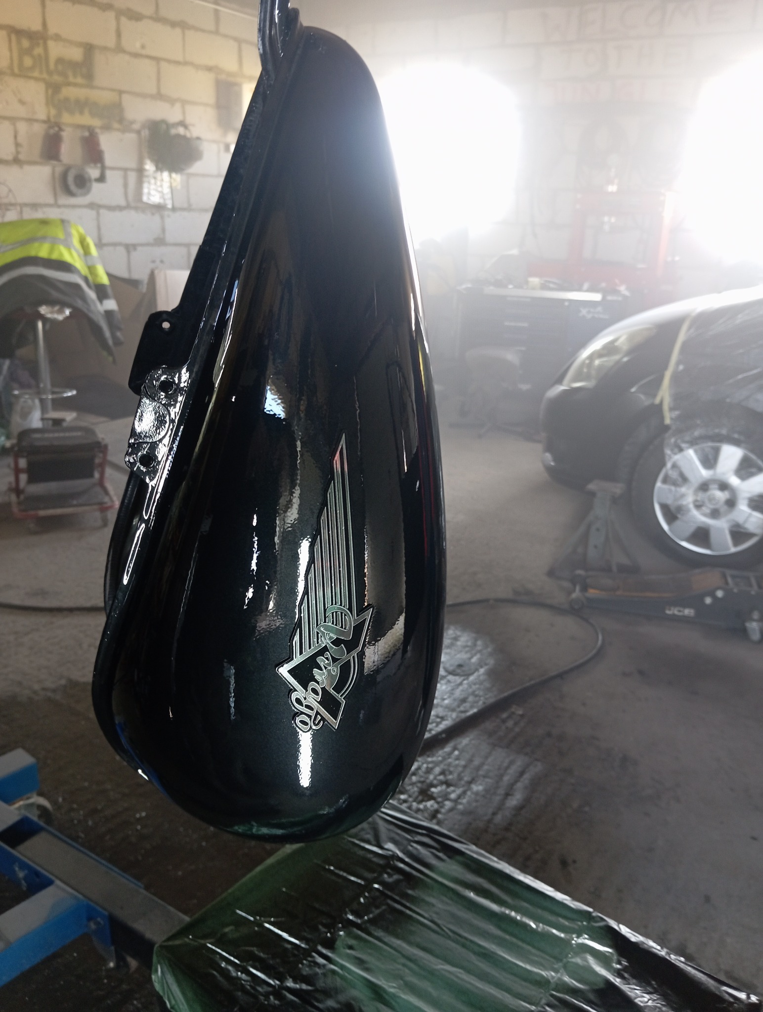 Błyszczący, czarny bak motocyklowy z logo, świeżo po lakierowaniu, zawieszony w warsztacie. W tle auto na podnośniku i narzędzia.