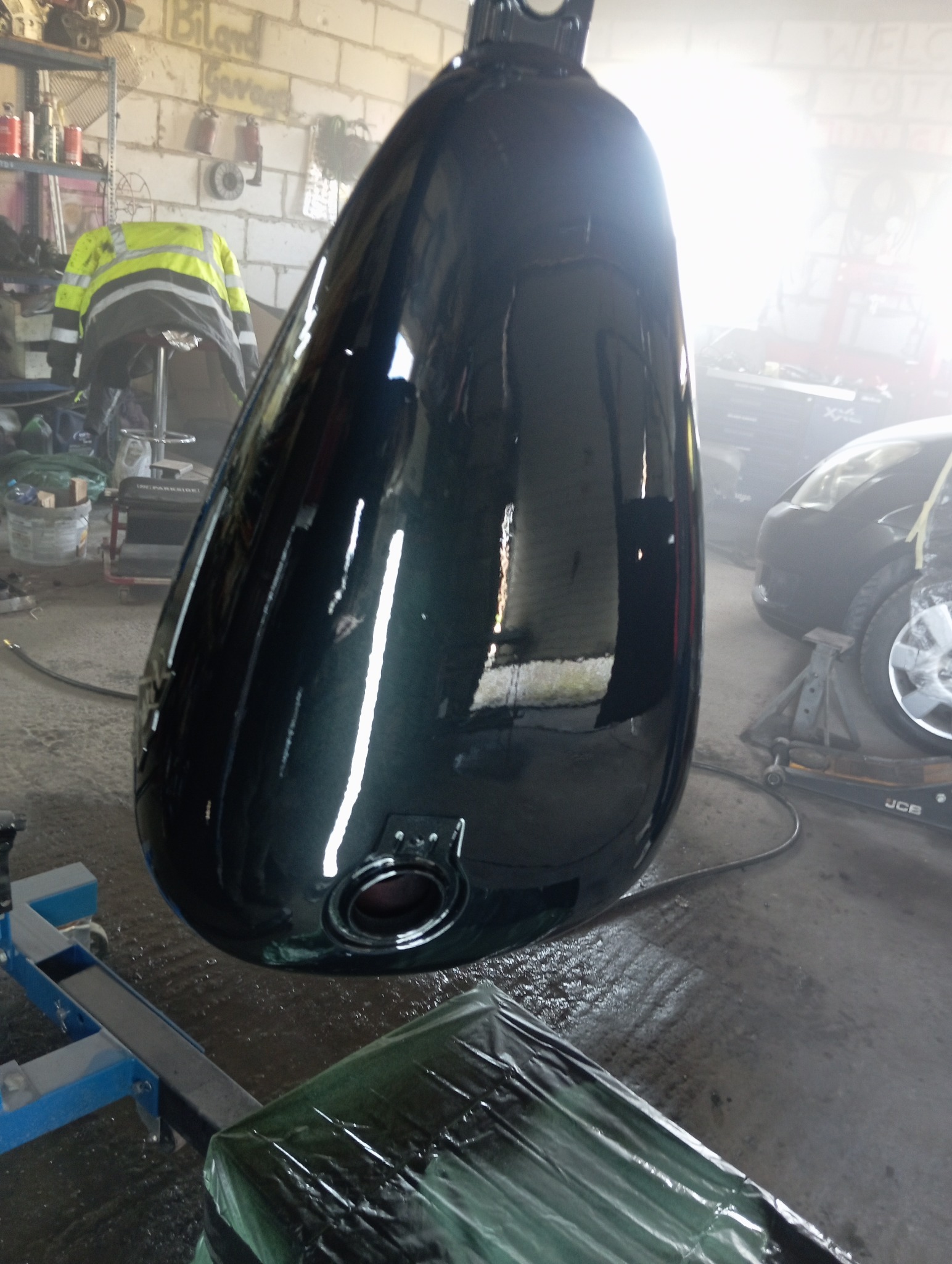 Błyszczący, ciemnozielony bak motocyklowy po lakierowaniu, zawieszony w warsztacie. Widoczne odbicia światła, fragment samochodu i narzędzia w tle.