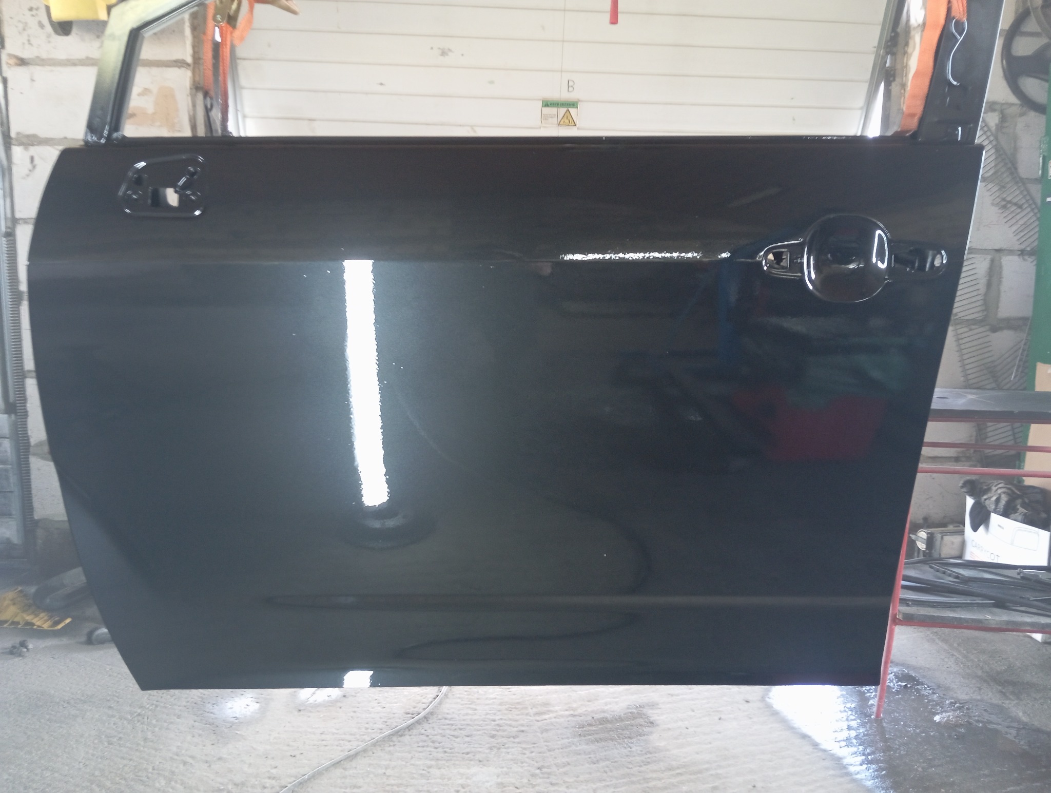 Polakierowane na czarno drzwi samochodowe z widoczną klamką, uchwytem i odblaskiem światła, w warsztacie z ceglanymi ścianami i narzędziami.