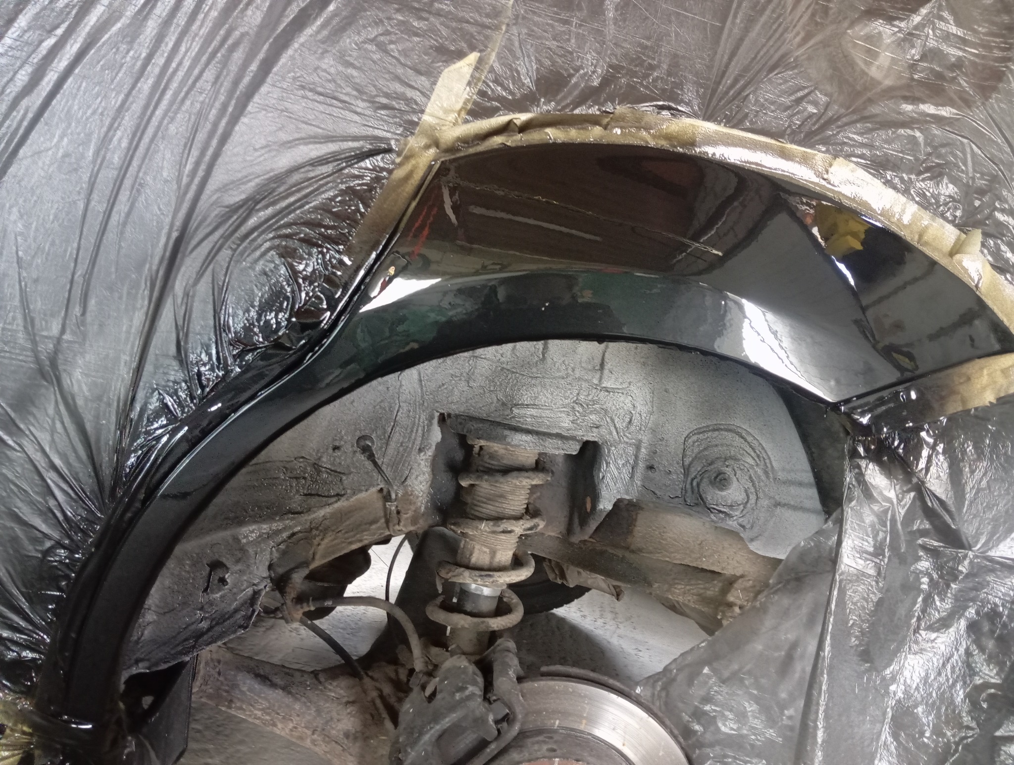 Zabezpieczone nadkole auta po nałożeniu warstwy ochronnej, widoczna sprężyna zawieszenia i elementy układu hamulcowego. Prace blacharsko-lakiernicze w toku.