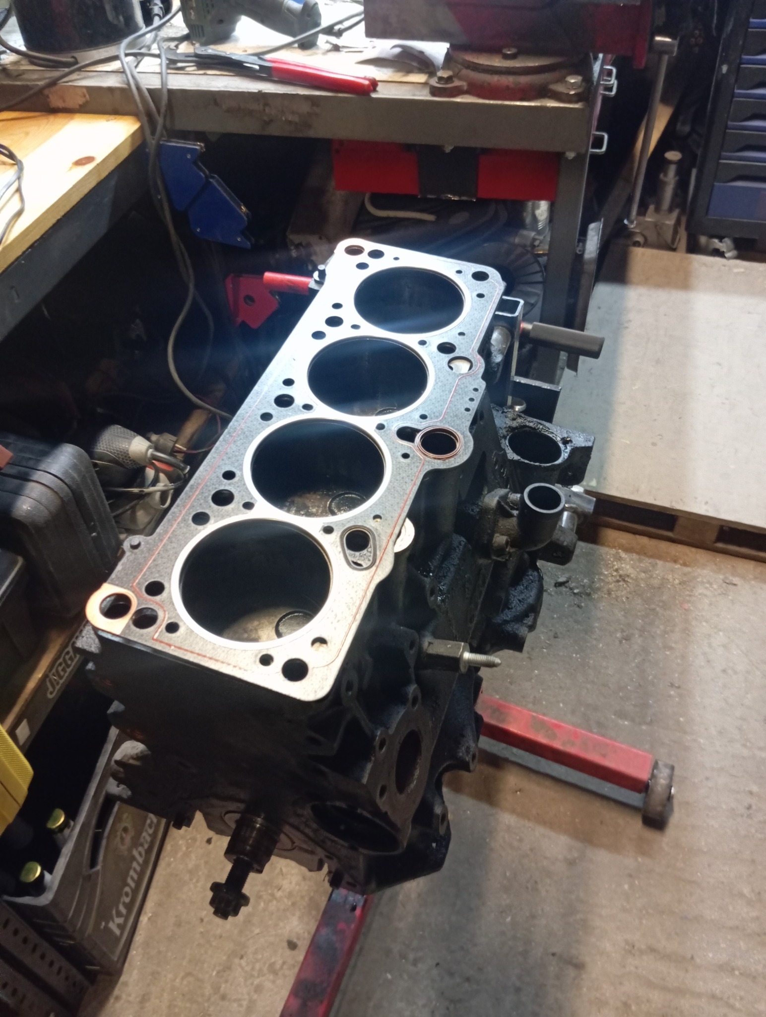 Częściowo zdemontowany silnik samochodowy z założoną uszczelką głowicy, stojący na czerwonym stojaku w warsztacie, ukazujący etap prac naprawczych.