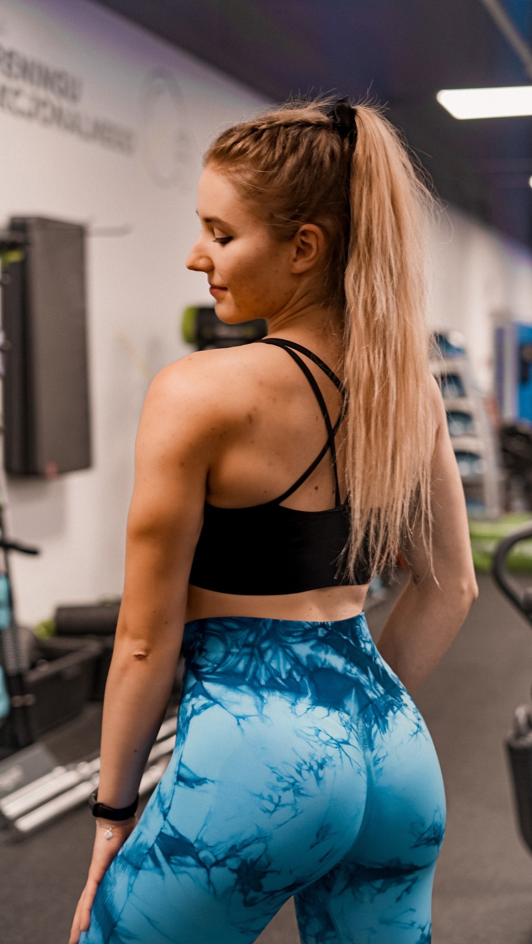 Kobieta w sportowym stroju (czarny top i niebieskie legginsy) stojąca tyłem w siłowni, z włosami spiętymi w wysoki kucyk i delikatnym makijażem.