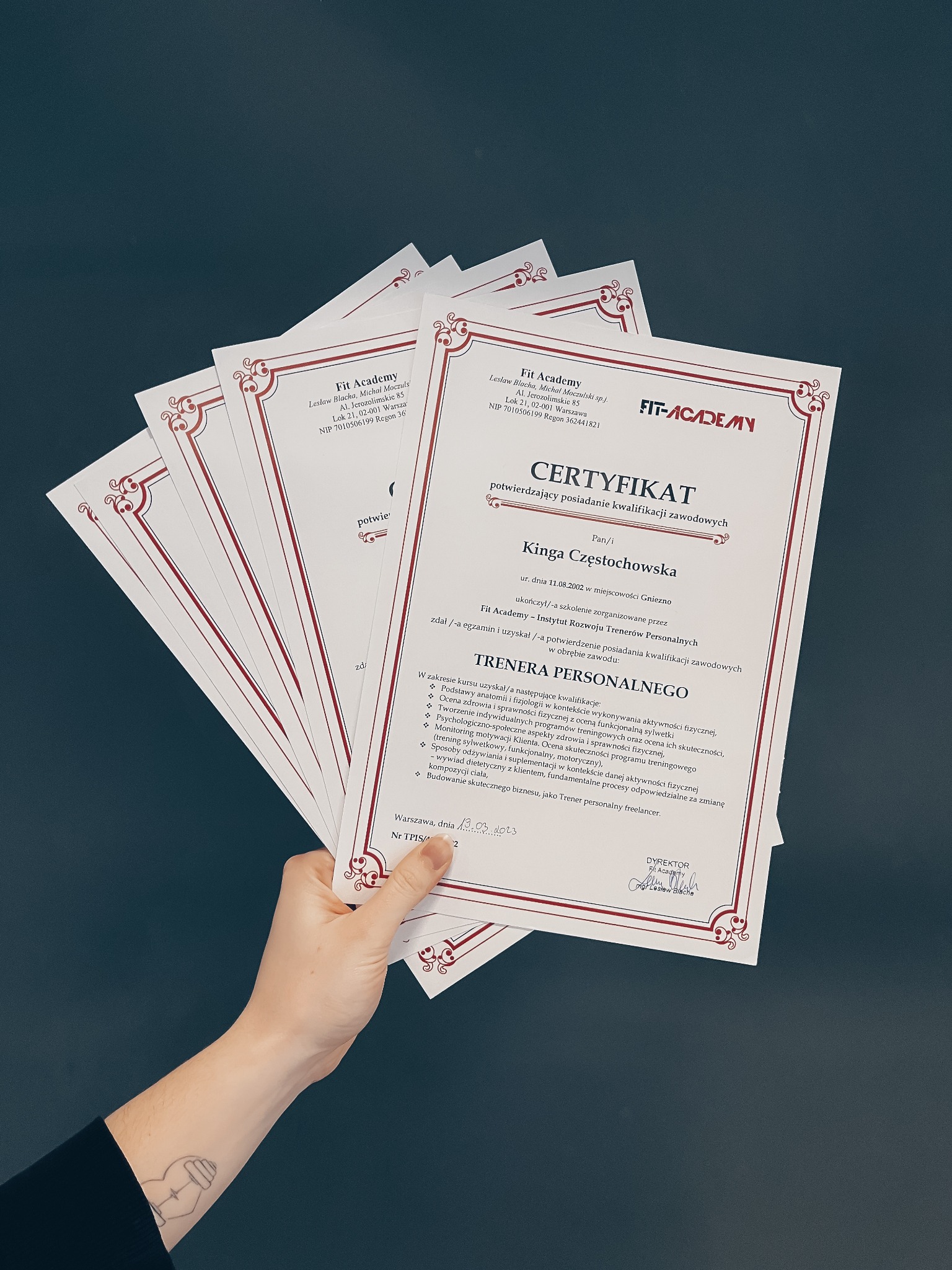 Dłoń trzymająca kilka certyfikatów ukończenia kursu na trenera personalnego z logo Fit Academy, widoczny tatuaż na przedramieniu.