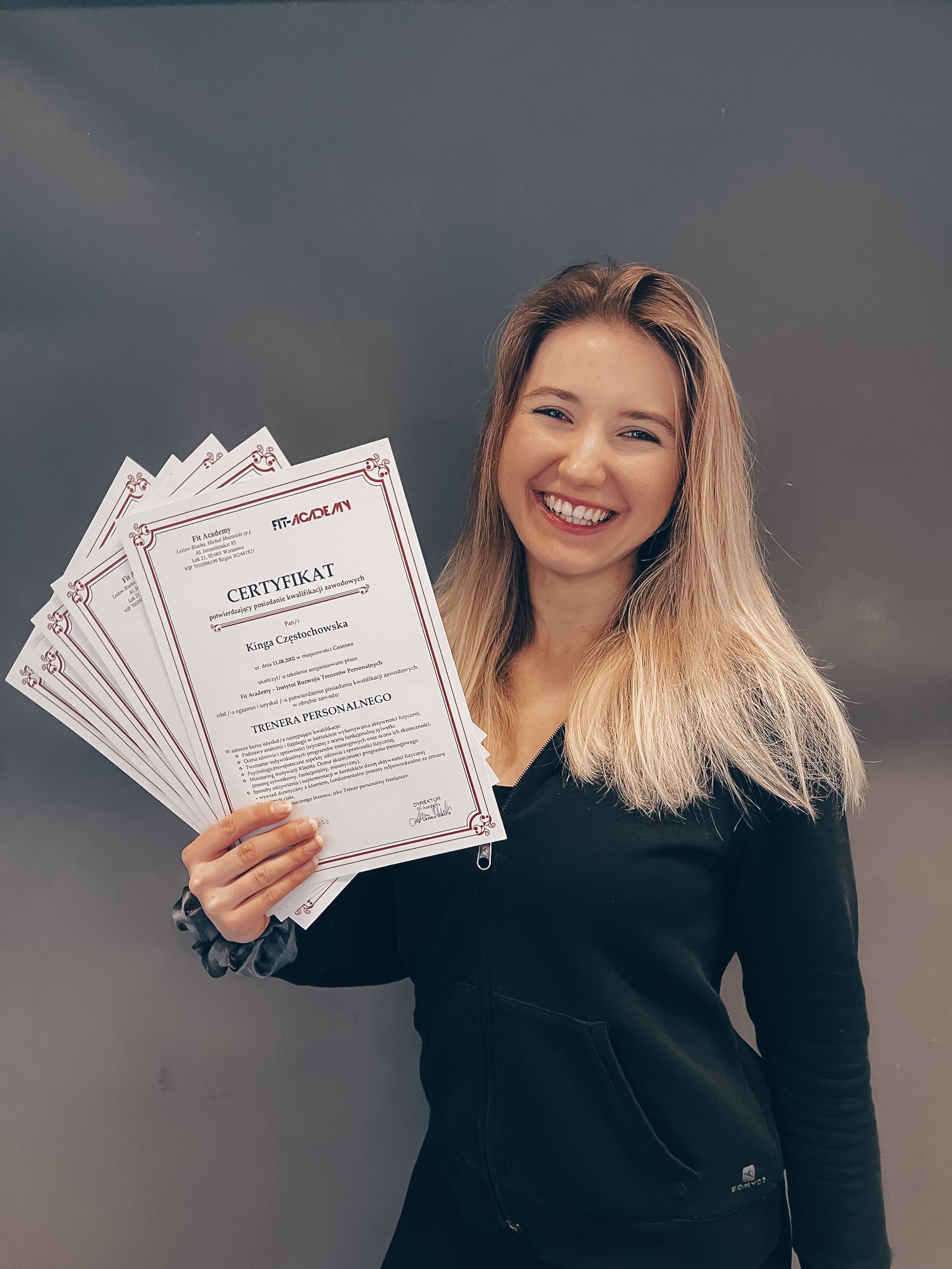 Uśmiechnięta blondynka w czarnej bluzie prezentuje kilka certyfikatów ukończenia kursu Trenera Personalnego Fit Academy na tle szarej ściany.