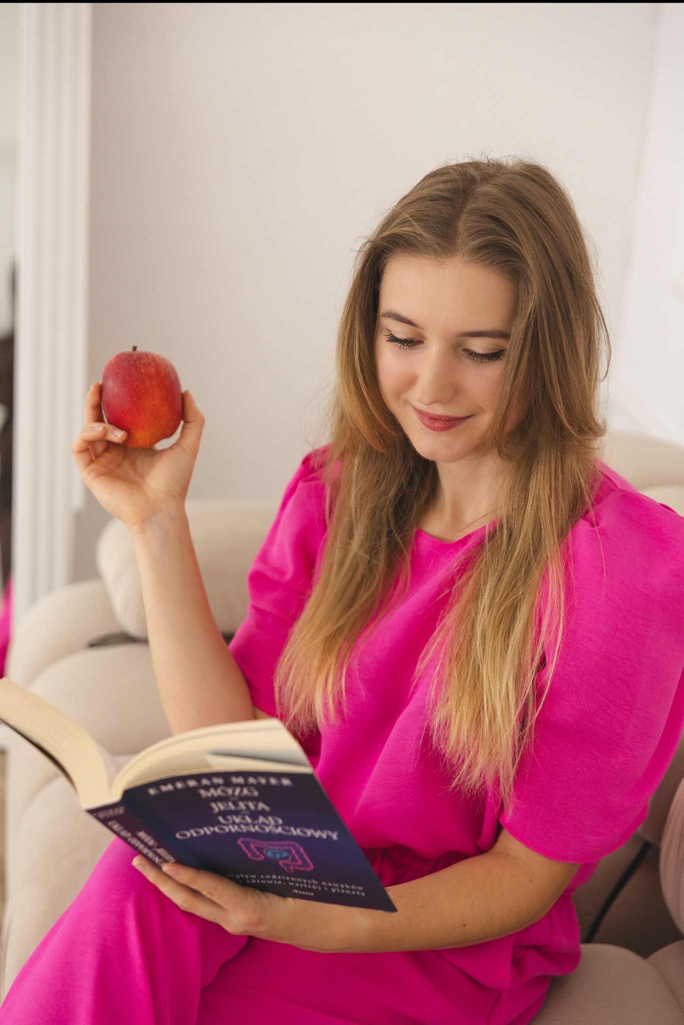 Kobieta w różowym stroju czyta książkę o zdrowiu, trzymając jabłko. Jasne wnętrze, naturalne światło, książka o jelitach i odporności. Zdrowy styl życia.
