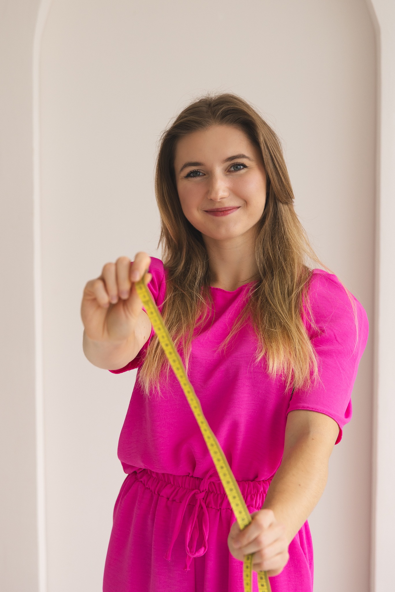 Uśmiechnięta blondynka w różowym stroju trzyma żółtą miarkę krawiecką, prezentując narzędzie pracy w jasnym wnętrzu. Pomiar obwodu ciała.