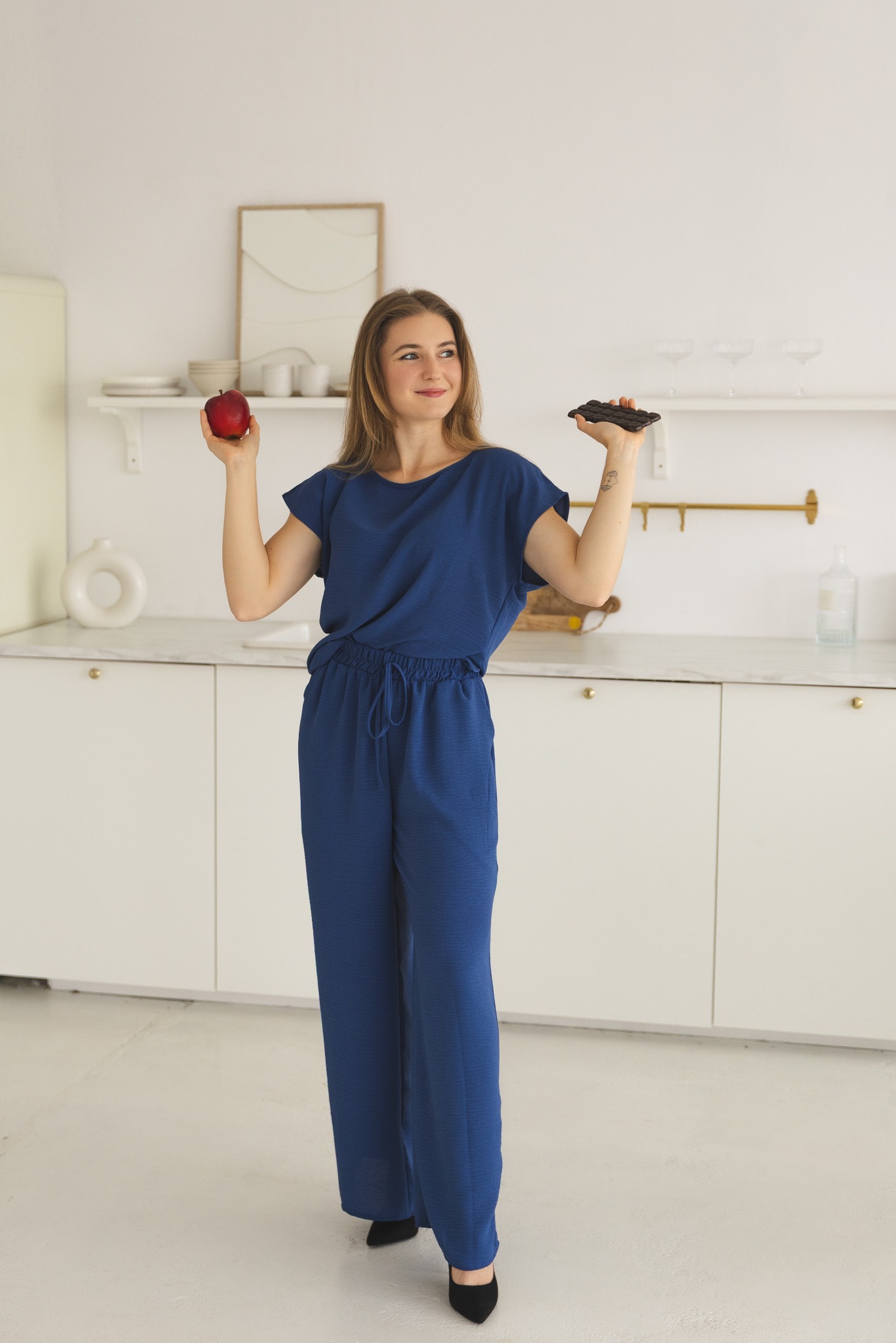 Uśmiechnięta kobieta w niebieskim stroju prezentuje jabłko i czekoladę na tle minimalistycznej kuchni. Kontrast zdrowego i niezdrowego jedzenia.