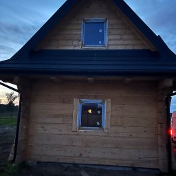 FENIX LINE SPÓŁKA Z OGRANICZONĄ ODPOWIEDZIALNOŚCIĄ - Dom z bali w trakcie montażu okien. Widoczne okna z folią ochronną, drewniana konstrukcja i czarny dach. Ujęcie z dołu, wieczorne światło.