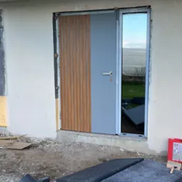 Nowe, szare drzwi zewnętrzne z drewnianymi panelami, zamontowane w surowym otworze budynku. Fragmenty styropianu na ziemi. Widoczne odblaski w szybie.