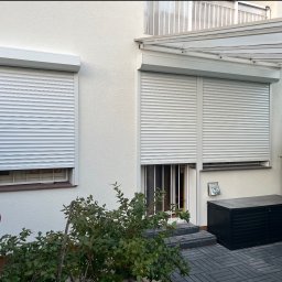 FENIX LINE SPÓŁKA Z OGRANICZONĄ ODPOWIEDZIALNOŚCIĄ - Biała fasada domu z dwoma oknami i drzwiami osłoniętymi roletami zewnętrznymi w kolorze białym. Nad drzwiami widoczny zadaszony ganek.