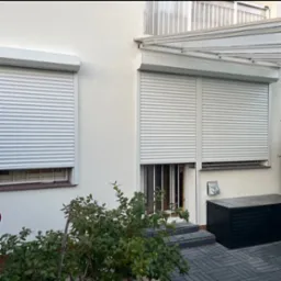 Biała fasada domu z dwoma oknami i drzwiami osłoniętymi roletami zewnętrznymi w kolorze białym. Nad drzwiami widoczny zadaszony ganek.