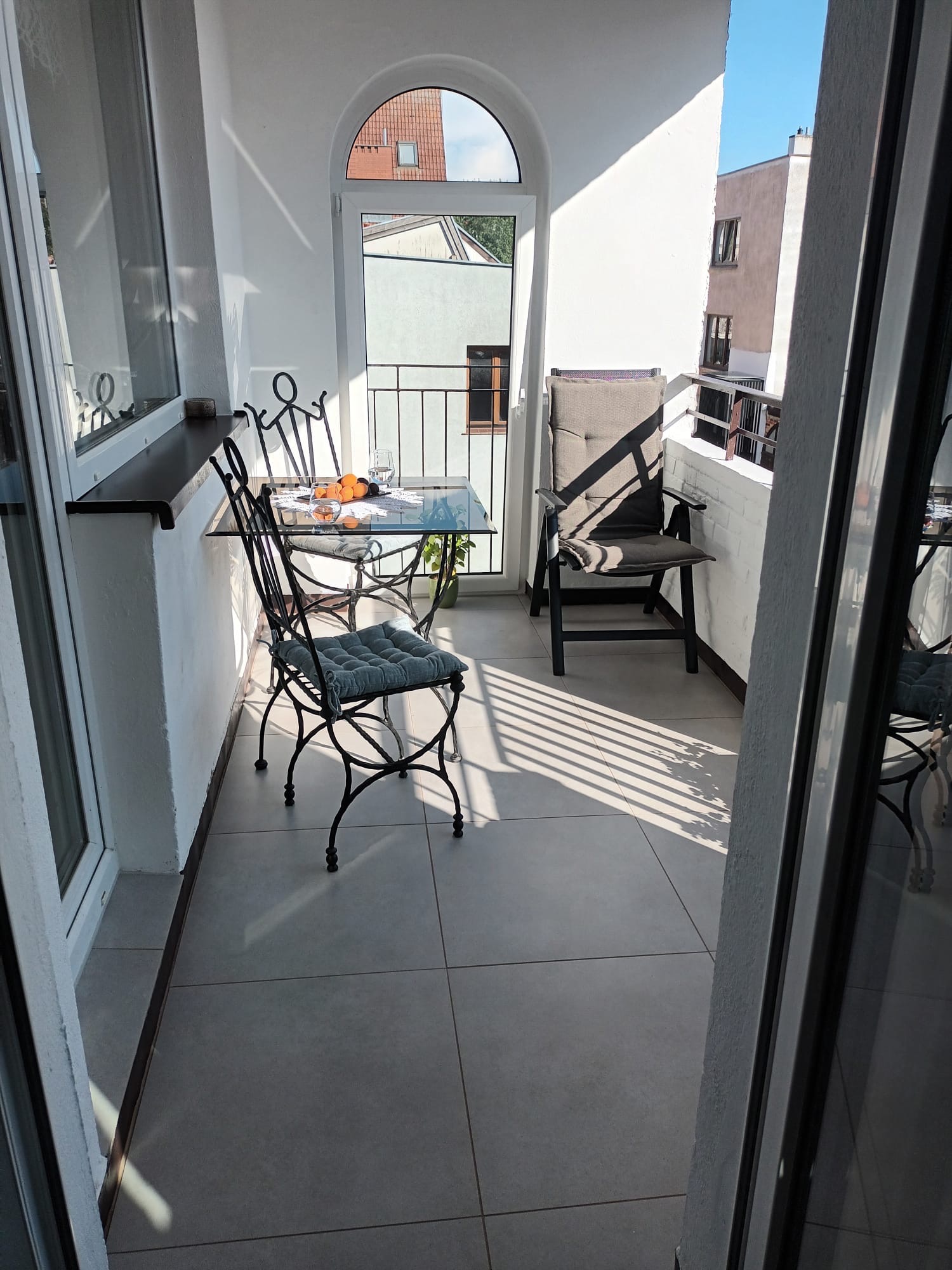 Jasny balkon z kutymi krzesłami i szklanym stolikiem, na którym leżą owoce. Widok na zewnątrz przez łukowe drzwi balkonowe.