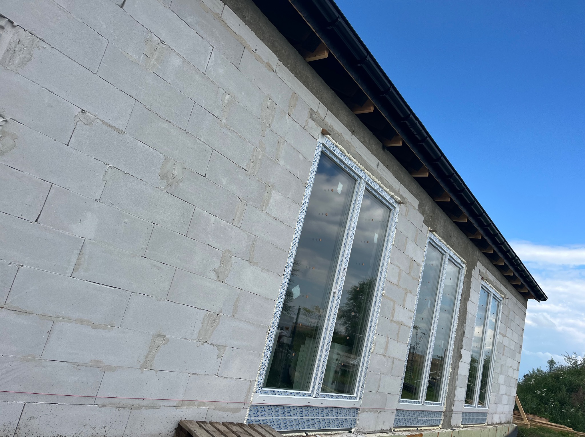 Montaż okien w surowym budynku z betonu komórkowego. Widoczne białe ramy okienne i niebieskie zabezpieczenia. Błękitne niebo w tle.