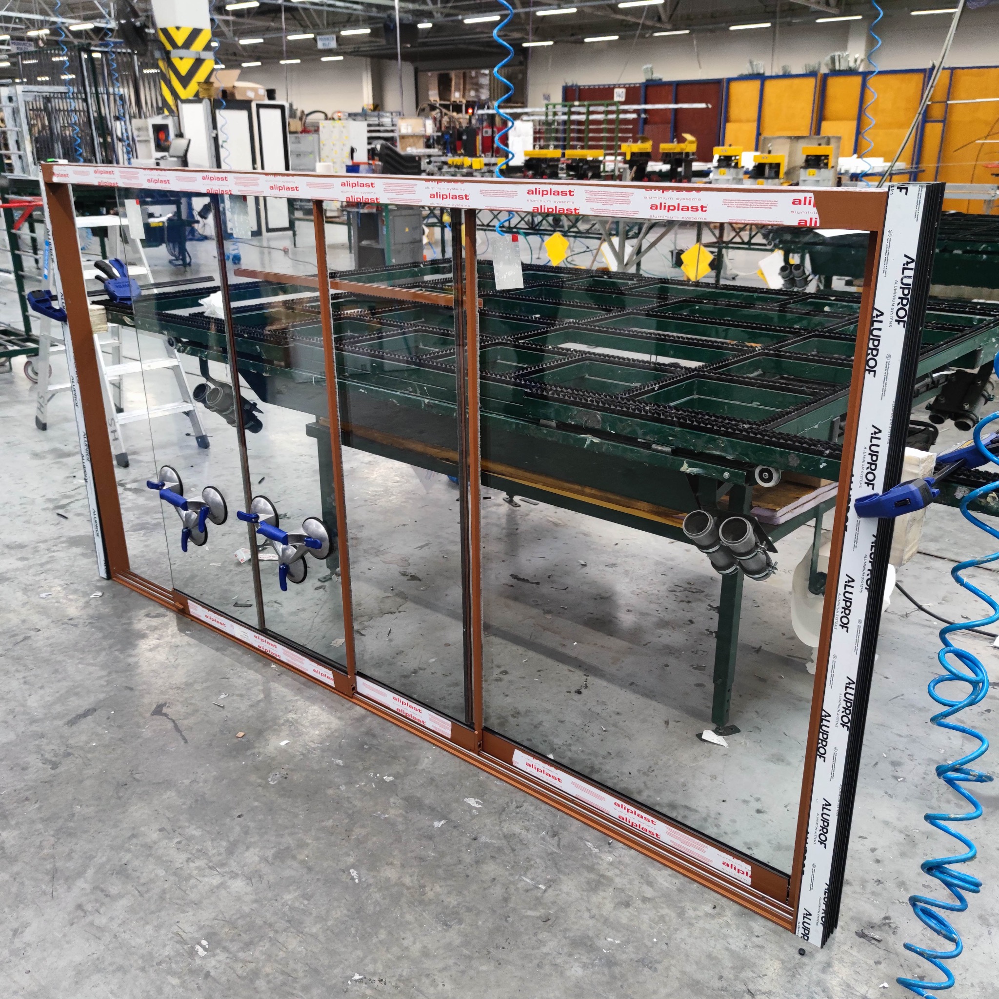 Brązowe okno przesuwne Aluplast i Aluprof z przyssawkami na szkle, ustawione na stole montażowym w hali produkcyjnej.