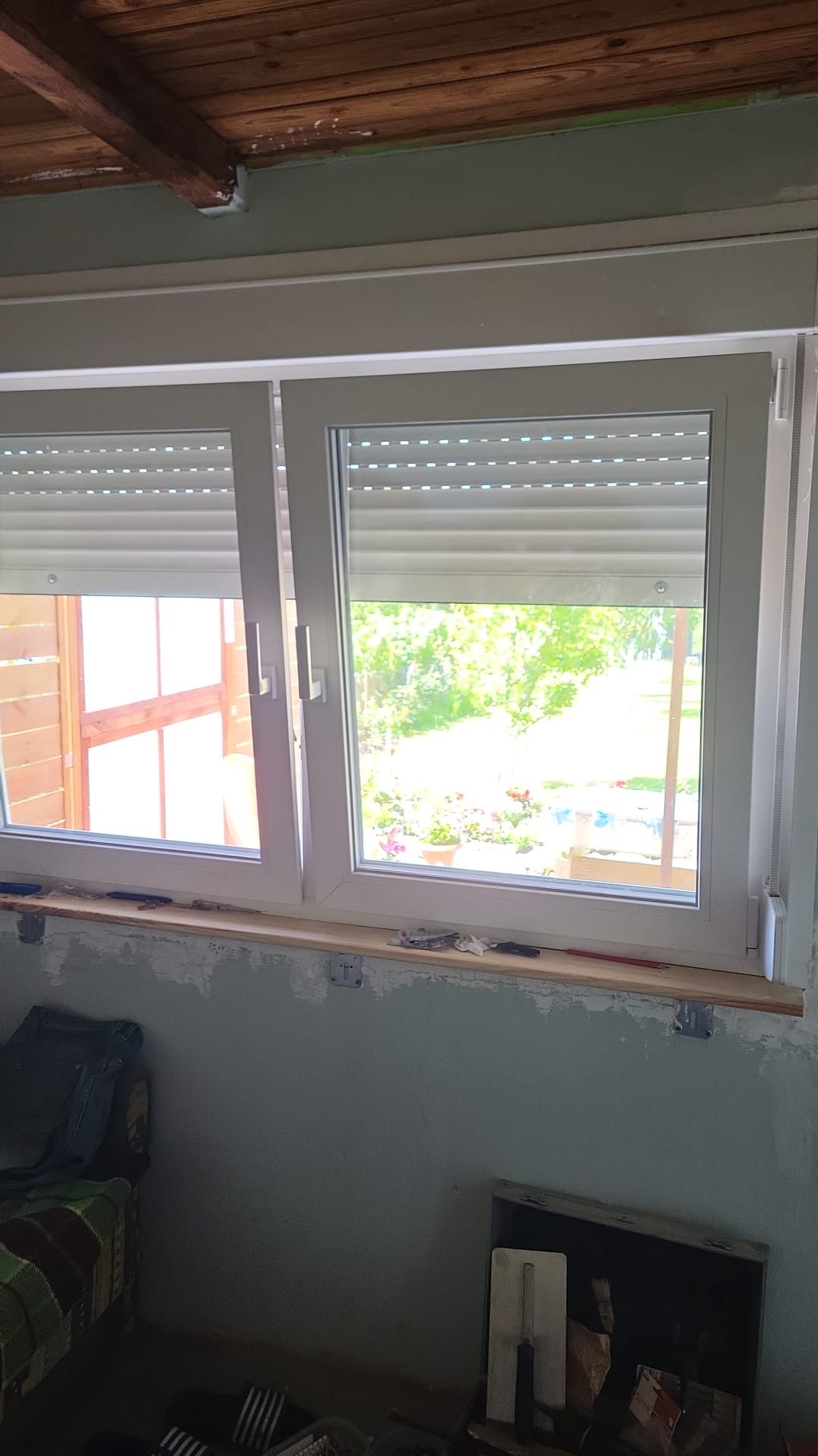 Białe okno PCV z roletami zewnętrznymi w trakcie montażu, widoczne narzędzia i fragment wnętrza z surową ścianą.