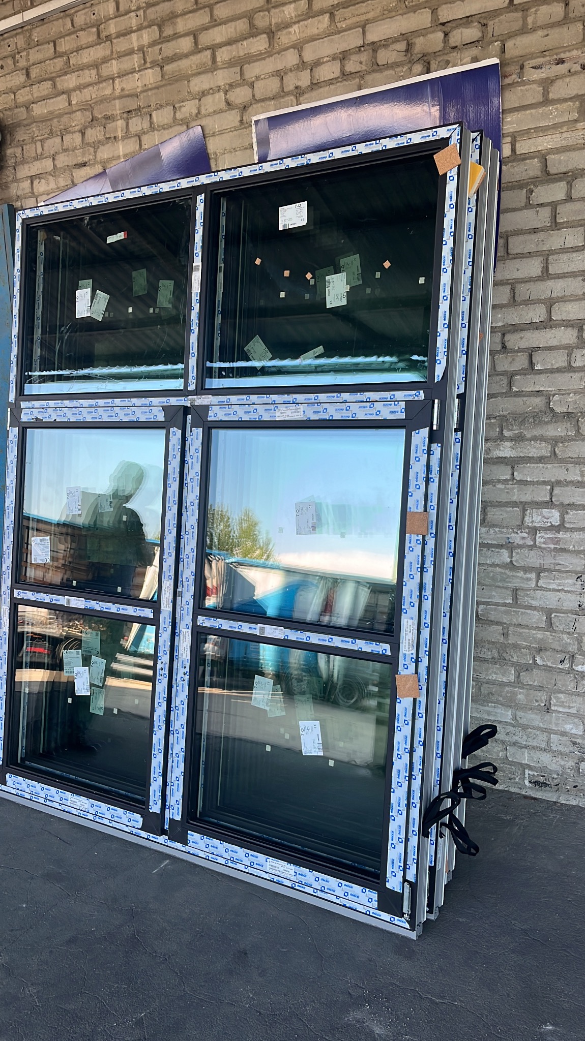Zestaw okien PCV z czarnymi ramami, zabezpieczonych niebieską folią, opartych o ceglaną ścianę. Widoczne etykiety na szybach i uchwyty do przenoszenia.